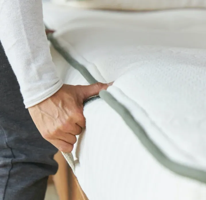A person’s hand lifts the edge of a white mattress topper on top of a mattress.