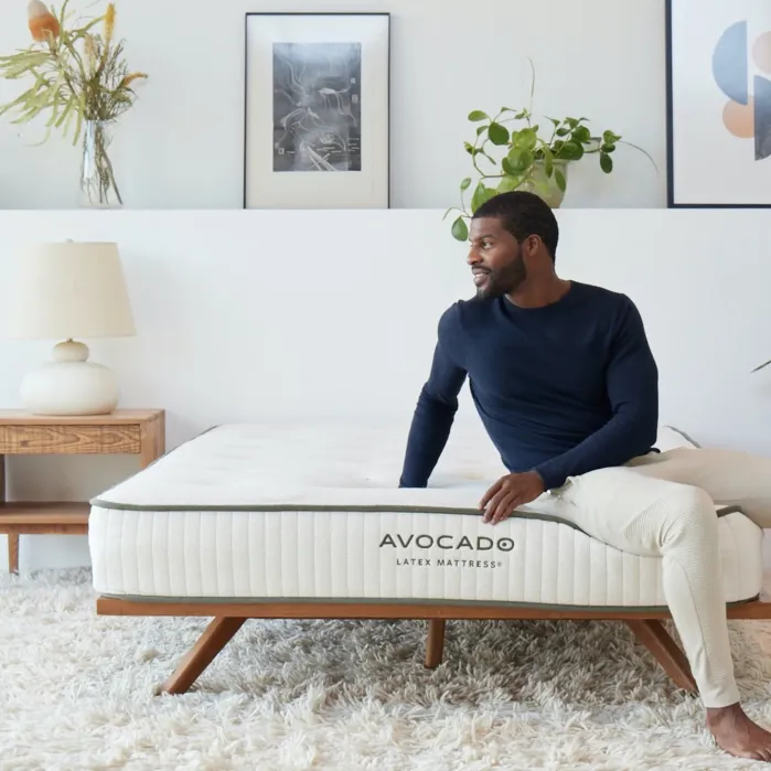 A man sits on the edge of an Avocado latex mattress in a modern, bright bedroom with plants and framed art on the wall.