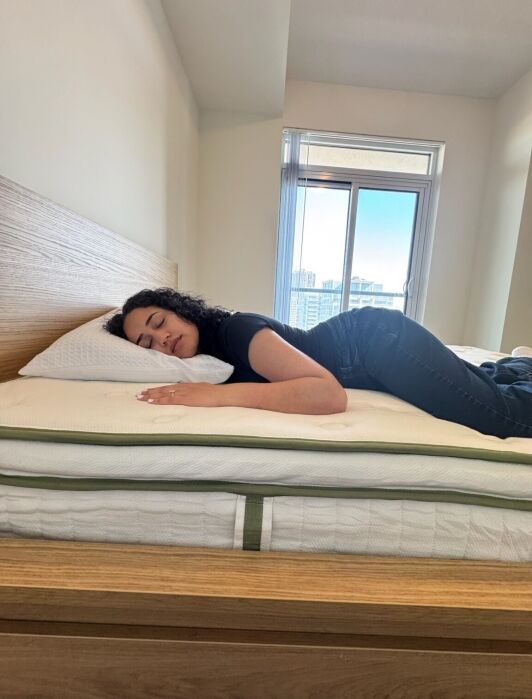 A woman lies on her stomach on a made bed with an organic mattress in a bright room, resting her head on a pillow with eyes closed. A window in the background reveals city buildings.