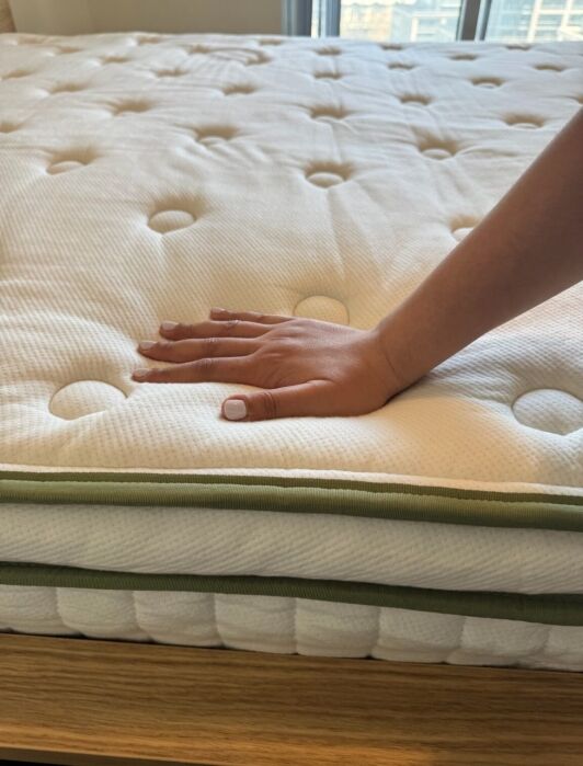 A hand presses down on a white, quilted organic mattress with green trim, highlighting its softness and texture.