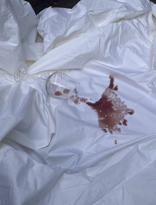 A clear wine glass lies on its side atop a white sheet, with red liquid spilled and spreading across the fabric.