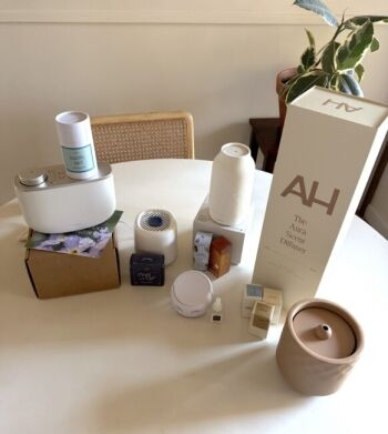 A variety of essential oil diffusers, scent cartridges, and fragrance boxes are arranged on a round white table with a chair and a plant in the background.