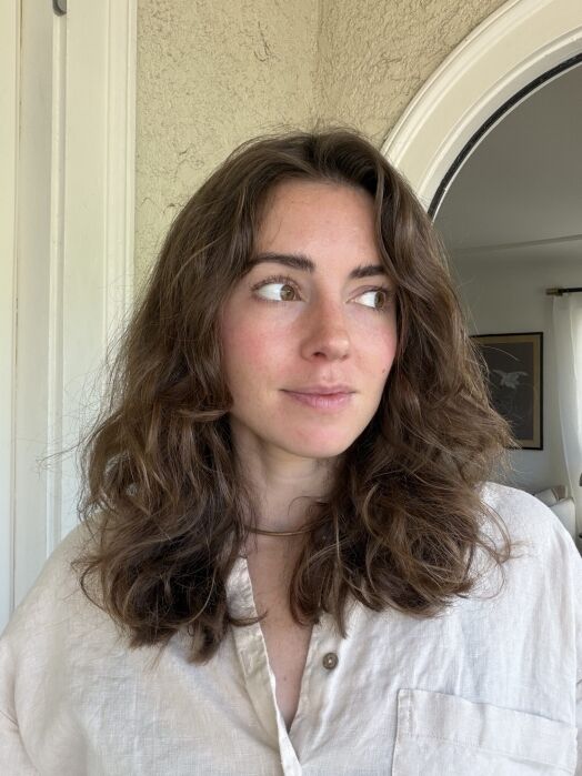 A woman with wavy brown hair and a beige shirt stands indoors, looking to her left with a neutral expression.
