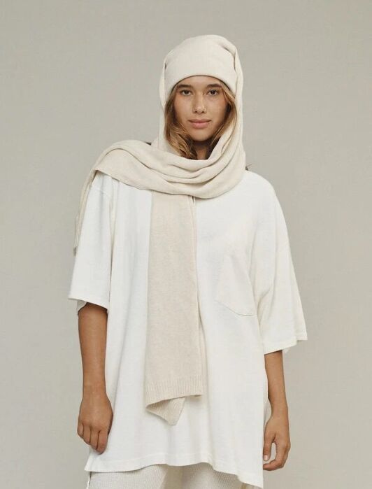 A woman stands against a plain background wearing an oversized white T-shirt, white knit pants, and a matching beige knit scarf and hat.