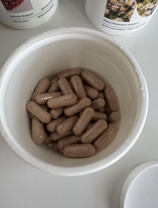 Open container filled with brown capsules, with two supplement bottles labeled "Immune Support" and "Sleep-Relaxation Support" partially visible in the background.