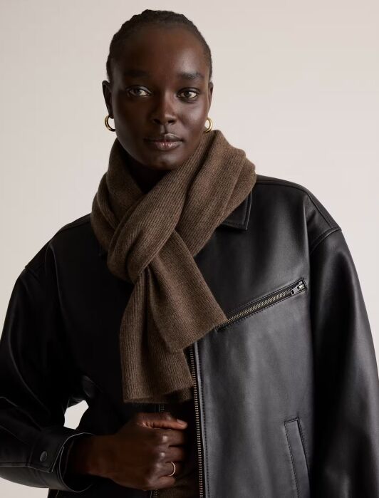 A person wearing a black leather jacket and a brown knit scarf, standing against a plain beige background.