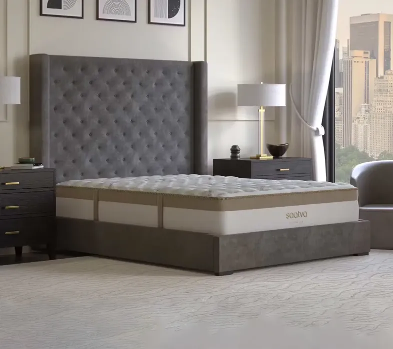 A bedroom with a large gray tufted headboard, two dark nightstands with lamps, a mattress on a platform bed, and a cityscape view through tall windows.