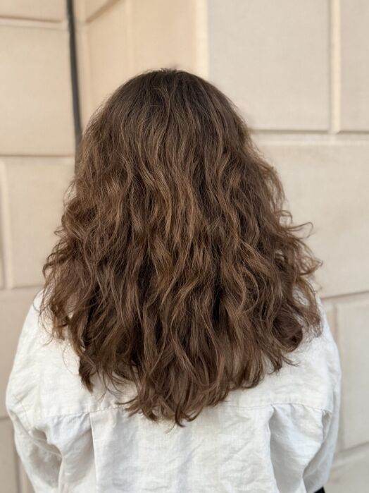 A person with medium-length, wavy brown hair is standing with their back to the camera, wearing a light-colored shirt in front of a beige wall.