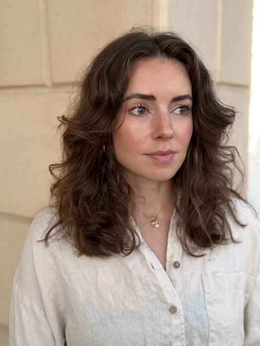 Woman with wavy brown hair wearing a light-colored button-up shirt stands in front of a beige textured wall, looking slightly to the side.