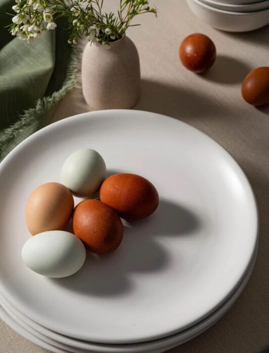 A white plate with four eggs of different colors sits on a table next to a small vase with white flowers and stacked bowls.