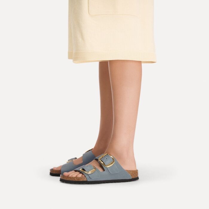 A person wearing a light beige skirt and gray double-strap sandals with gold buckles, standing against a plain white background.