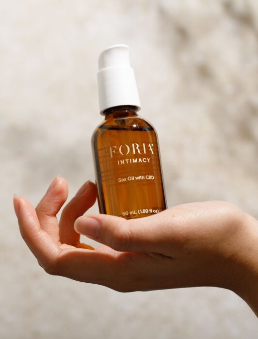 A hand holds a bottle of FORIA Intimacy Sex Oil with CBD against a blurred, neutral background.