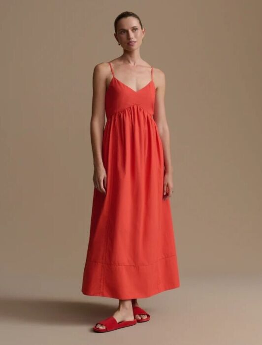 A woman stands against a plain beige background, wearing a sleeveless, ankle-length red dress with spaghetti straps and matching red slides.