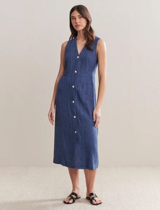 A woman stands wearing a sleeveless, button-down, blue midi dress with a V-neck, paired with black sandals, against a plain background.