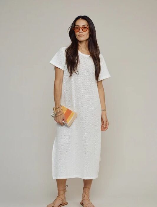 A woman wearing a white short-sleeve midi dress, tan sandals, orange sunglasses, and holding a striped clutch stands against a plain background.