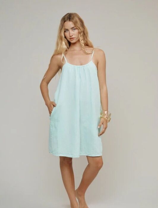 A woman with long blonde hair wears a loose, light blue sleeveless dress with thin straps and stands barefoot against a plain light background.