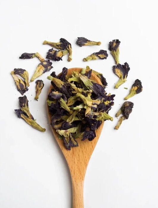A wooden spoon holds dried purple and green butterfly pea flowers, with additional flowers scattered on a white background.