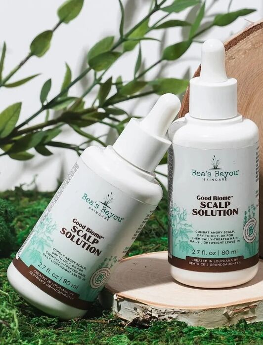 Two bottles of Bea's Bayou Skincare Good Biome Scalp Solution are displayed on a mossy surface with green leaves in the background.