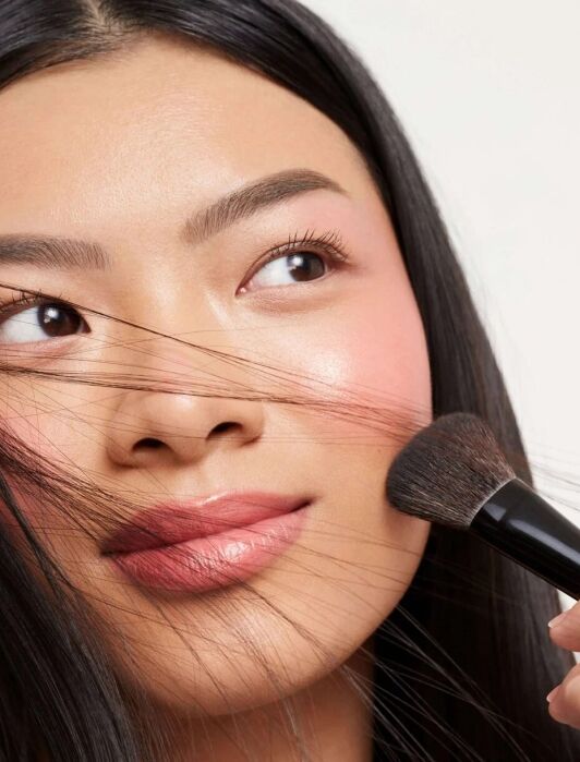 A person with long dark hair applies blush to their cheek with a makeup brush; some hair strands are across their face.