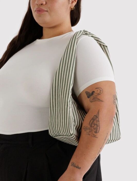 A person wearing a white short-sleeve top, black pants, and a green striped bag over their shoulder, with visible tattoos on their arm.
