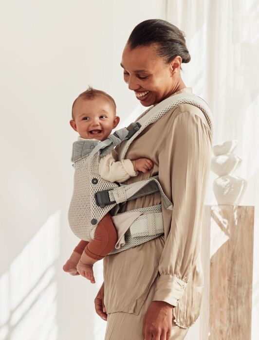 An adult wearing a light-colored outfit carries a smiling baby in a front-facing baby carrier. The scene is indoors with soft natural light.