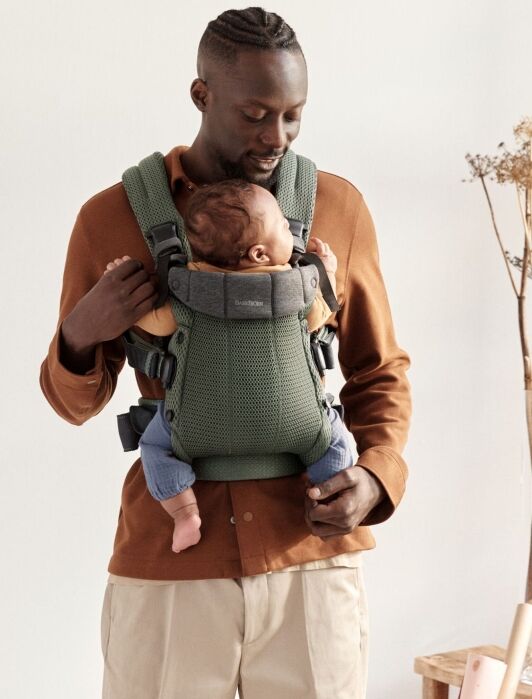 A man wearing a brown shirt carries a baby in a green front-facing baby carrier while standing indoors.