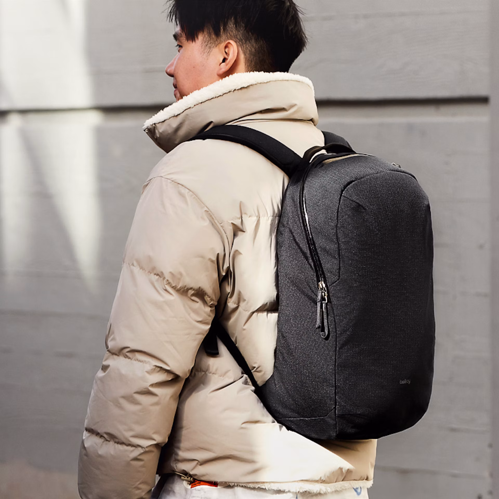Person wearing a beige puffer jacket and carrying a large black backpack stands in front of a light gray wall.
