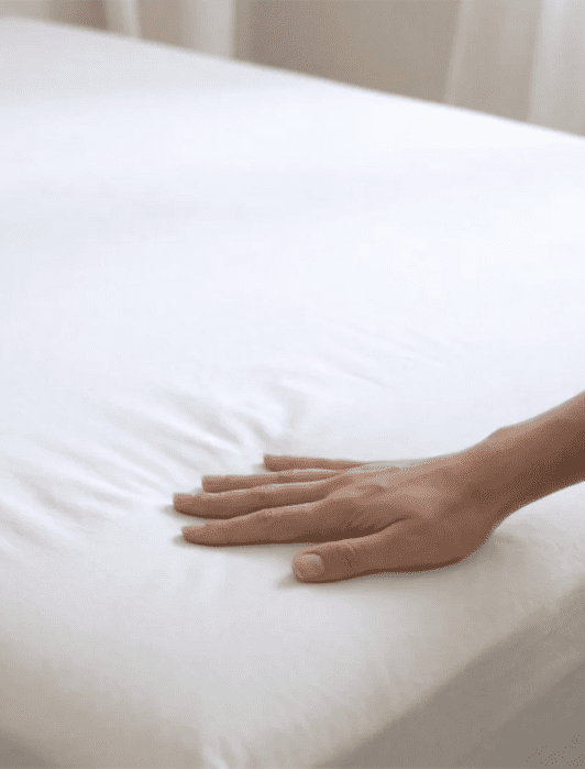 A hand pressing down on a neatly made bed with a white fitted sheet.