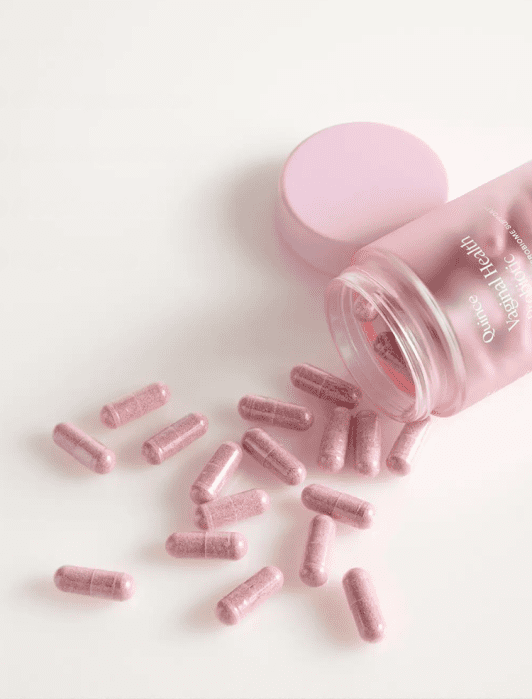 A pink bottle labeled “Women’s Health” lies on its side with pink capsules spilling out; the lid is nearby on a white surface.