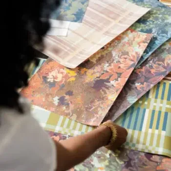 A person arranges sheets of patterned fabric samples featuring floral and plaid designs in various colors on a table.