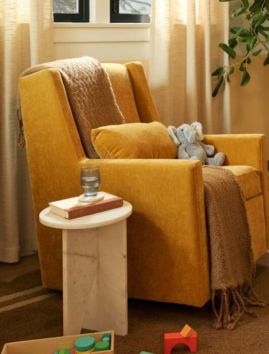 A yellow armchair with a brown throw and stuffed toys next to a round side table and a wooden crib in a cozy, softly lit nursery.