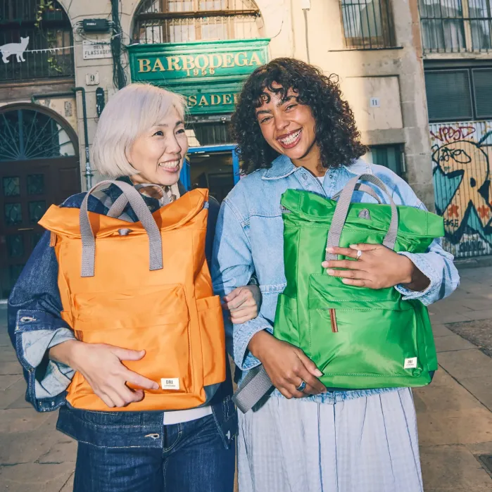 Two women standing outside, smiling and holding brightly colored backpacks, one orange and one green, in front of a building with a green sign that reads “BARBODEGA.”.