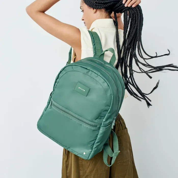 Person with long braids wearing a sleeveless shirt holds a large green backpack over one shoulder, facing away from the camera against a plain background.