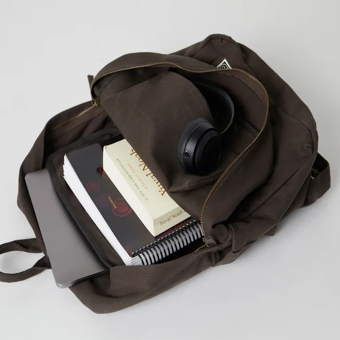 A brown backpack containing a laptop, books, a spiral notebook, a pencil pouch, and black over-ear headphones on a white surface.