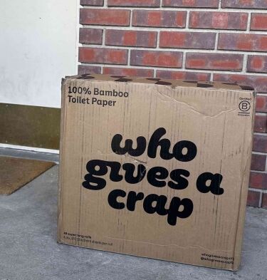 A cardboard box labeled "who gives a crap" 100% bamboo toilet paper sits on a concrete porch near a red brick wall and a doormat, showcasing the eco-friendly ethos of Who Gives A Crap.