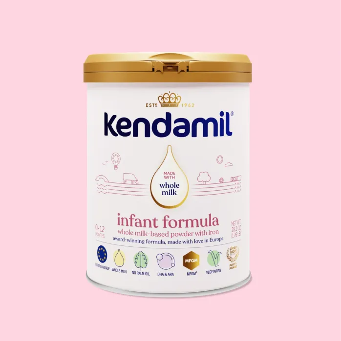 A can of Kendamil infant formula with a gold lid, labeled “whole milk-based powder with iron” for ages 0-12 months, displayed on a pink background.