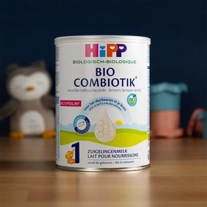 A can of HiPP Bio Combiotik infant formula sits on a wooden surface with a blurred owl toy and other objects in the background.