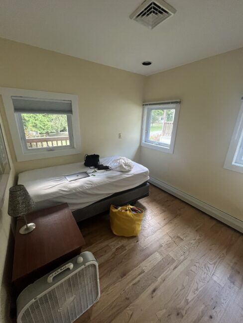A small bedroom with a single bed, a side table with a lamp, a floor fan, a yellow bag, and two windows with views of greenery outside.