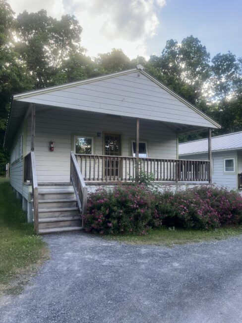 A small, light-colored cabin with a front porch, wooden railing, and stairs, surrounded by pink flowering bushes and trees in the background.