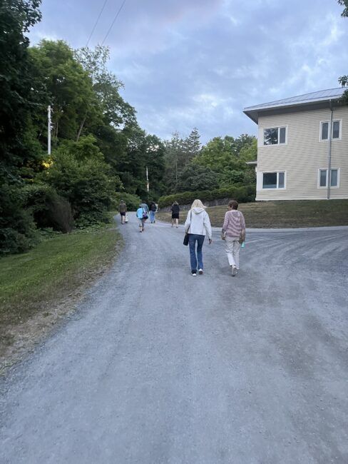 A group of people walk up a wide gravel road bordered by trees and a beige building under a partly cloudy sky.