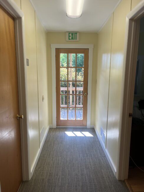 A narrow hallway with yellow walls, two closed wooden doors on each side, and a glass door at the end leading outside with an exit sign above it. Sunlight shines through the glass.