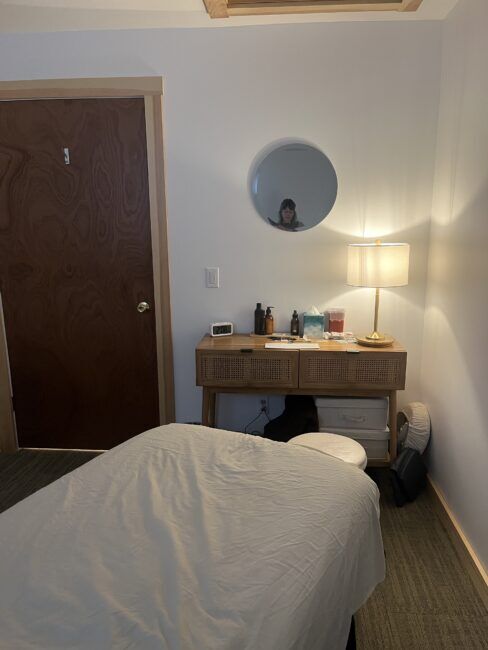 A small room with a massage table, a wooden dresser holding bottles and tissues, a lamp, and a round mirror reflecting a person.