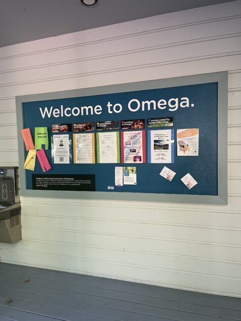 A bulletin board labeled "Welcome to Omega" displays various flyers, notices, and colored paper on a wooden wall near a water fountain.