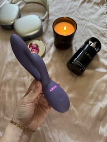 A hand holds a purple LELO vibrator above a bed, with wireless headphones, a lit candle, a bottle, and a ring in the background.