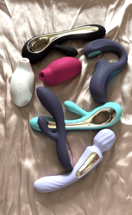 Six assorted silicone vibrators in various shapes and colors, including purple, teal, white, pink, and black, are arranged on a satin fabric background.