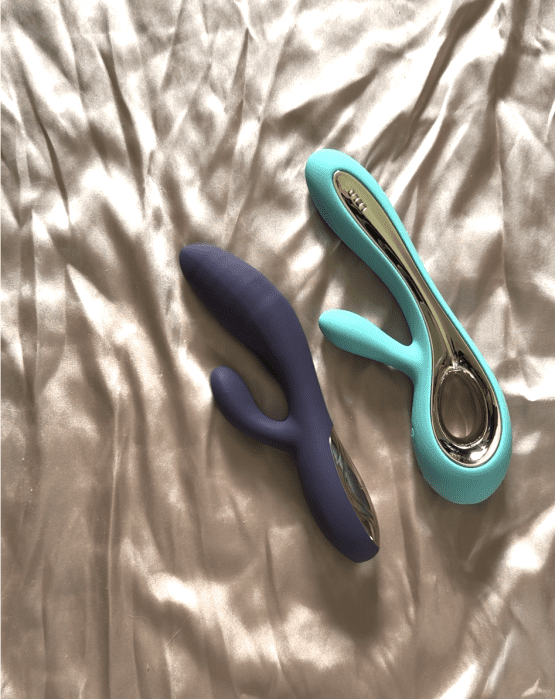 Two silicone rabbit vibrators, one purple and one blue with metallic accents, are placed on a beige satin bedsheet.