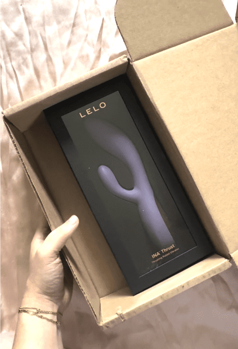 A hand holds an open cardboard box containing a LELO INIA Thrust personal massager in its packaging.