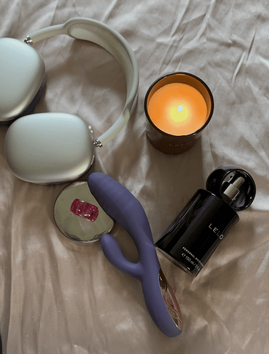 A pair of over-ear headphones, a lit candle, a bottle of LELO perfume, a purple vibrator, and a small pink accessory on a soft beige surface.