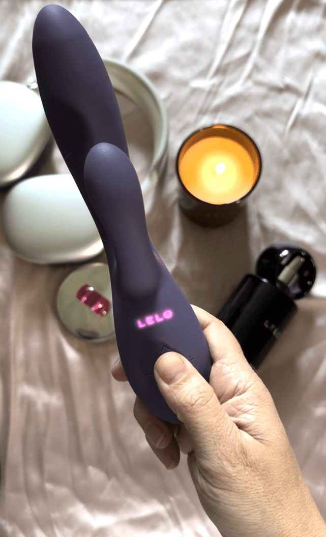 A hand holding a purple LELO vibrator above a bed, with a lit candle, black bottle, pink lipstick, and white headphones nearby.