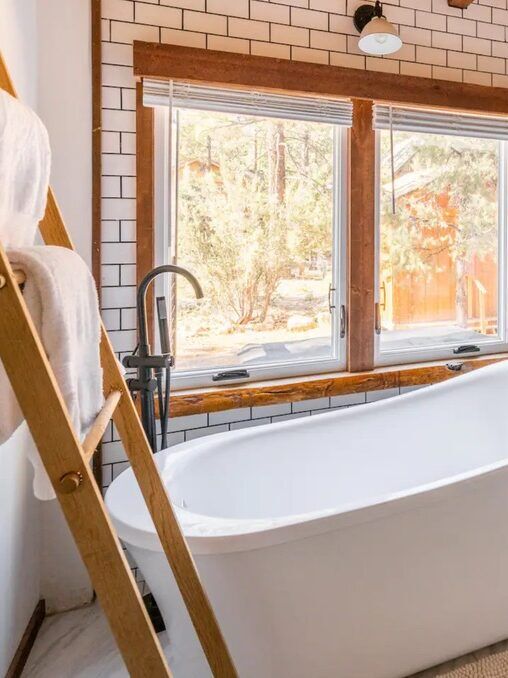 Bathroom with a white freestanding tub, wall-mounted sink, round mirror, ladder towel rack, and large windows overlooking trees. White subway tiles and wood accents throughout.